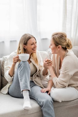 Moda kıyafetleri içinde, kanepede oturmuş el ele çay içip kaynaşan iki sevimli kız kardeş.
