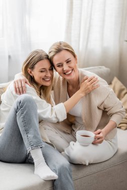 İki güzel kız kardeş şık giyinmiş, kanepede oturup sarılıyorlar, aile bağı kuruyorlar.