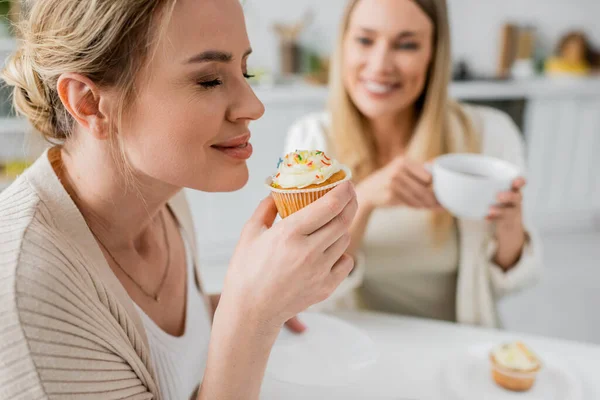 stock image close up two trendy sisters sitting at table drinking tea and eating cupcakes, closed eyes, bonding