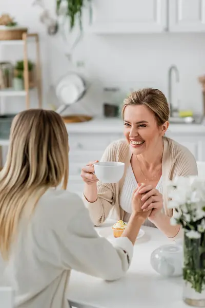 stock image classy smiley blonde sisters in pastel cardigans holding hands and drinking tea, family bonding