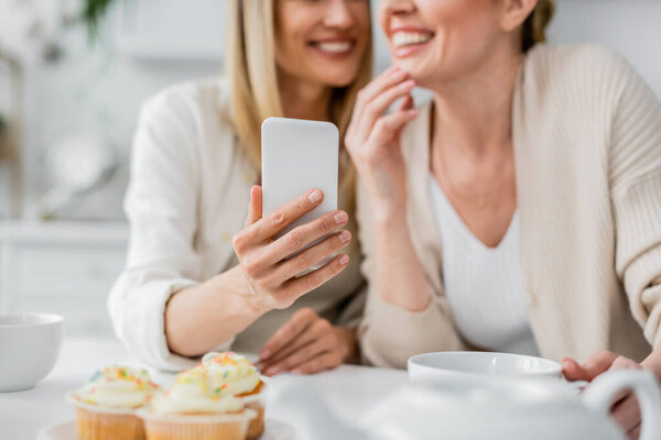 cropped view of two attractive sisters in cardigans making selfie on kitchen backdrop, bonding