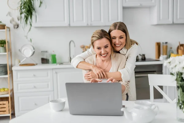 attractive blonde sisters looking at laptop smiling and hugging from behind, family bonding