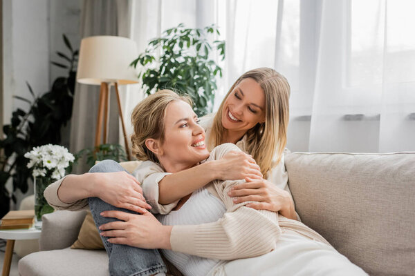 two lovely beautiful sisters hugging warmly on sofa looking at each other, togetherness, bonding