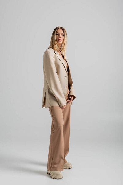 stylish beautiful woman in beige and brown suit looking at camera posing on grey backdrop, fashion