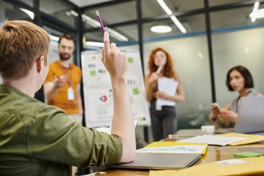 Toplantı odasında takım konuşmasına soru sorarken iş adamı kalem kaldırıyor.