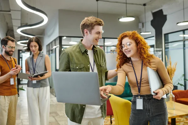 红头发女人指着同事和经理们正在谈论办公室背景的笔记本电脑 — 图库照片