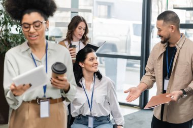 smiling asian man holding folder and discussing project with businesswoman near female colleagues clipart