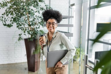 Modern ofiste elinde kağıt bardak ve dizüstü bilgisayarla mutlu bir Afrikalı Amerikalı iş kadını.