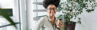 happy african american businesswoman in glasses holding paper cup with coffee in office, banner clipart