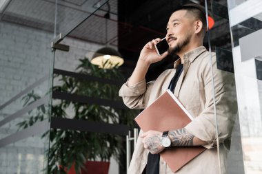 Dövme klasörlü mutlu Asyalı işadamı ve cam kapının yanında akıllı telefondan konuşuyor.