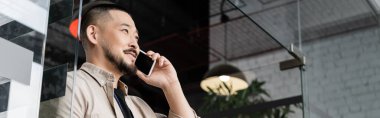 Mutlu Asyalı bir adam, ofisteki cam kapının yanındaki akıllı telefondan iş görüşmesi yapıyor.