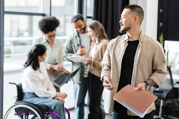 весёлый азиат держит смартфон и папку рядом с мультикультурной командой на размытом фоне