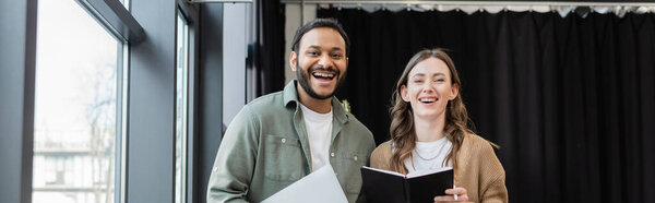 радостная женщина с ноутбуком и индийский мужчина с ноутбуком, смотрящий на камеру в офисе, коворкинг баннер