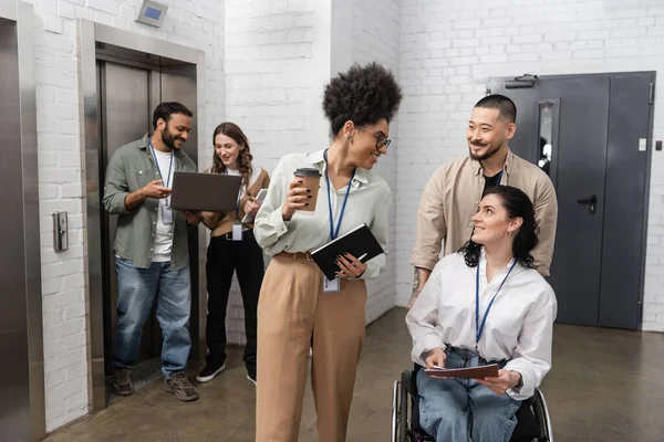 stock image asian man pushing wheelchair of disabled businesswoman and chatting with african american colleague