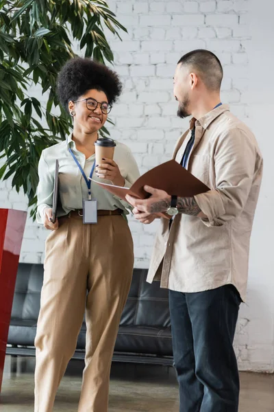 stock image cheerful african american woman holding coffee to go and chatting with asian coworker during break