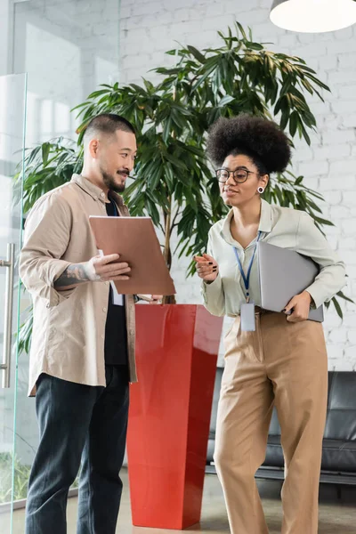 asian man showing folder with startup plan to african american woman with laptop in office hall