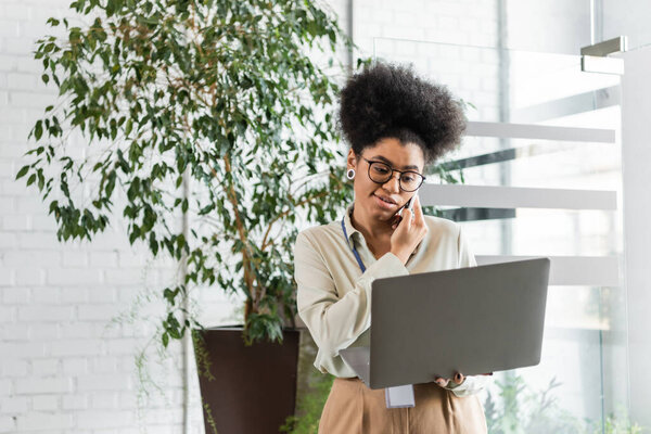 african american businesswoman in glasses holding laptop and talking on smartphone, multitasking
