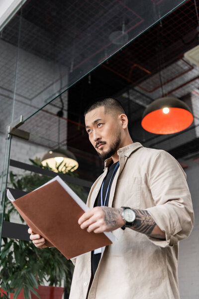 serious asian team leader with tattoo holding startup plan inside of folder near glass door
