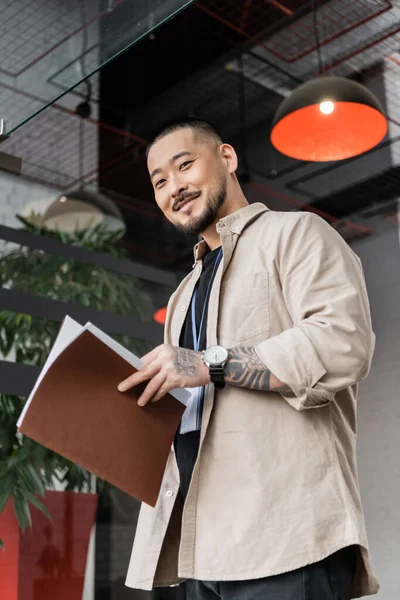 stock image cheerful asian office worker with tattoo holding folder and looking at camera near glass door