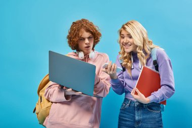 Akıllı telefonlu heyecanlı genç kız mavi ekrandaki düşünceli kızıl öğrencinin yanında dizüstü bilgisayarı işaret ediyor.
