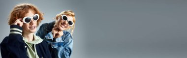 happy teen friends in stylish casual attire and sunglasses posing and smiling on grey, banner clipart