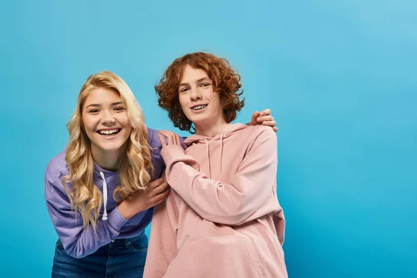 stock image blonde teenage girl hugging redhead boyfriend and laughing at camera on blue, happiness and unity