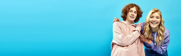 cheerful and trendy teenagers in hoodies embracing and looking at camera on blue, banner