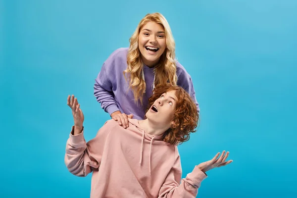 stock image excited blonde teen girl having fun and laughing at camera near surprised redhead boyfriend on blue