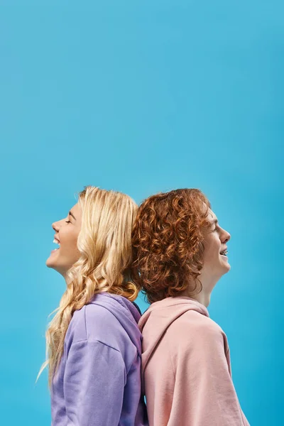side view of excited blonde girl with redhead friend laughing with closed eyes back to back on blue