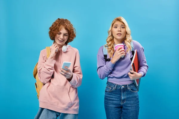 stock image nervous teen girl with paper cup looking at camera near positive friend using smartphone on blue