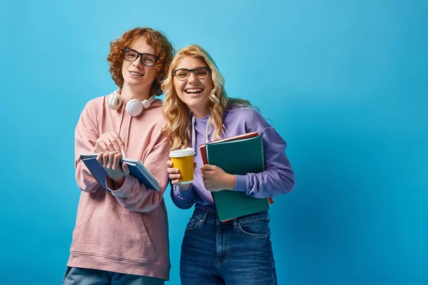 excited teen students in eyeglasses holding notebooks and coffee to go and smiling at camera on blue