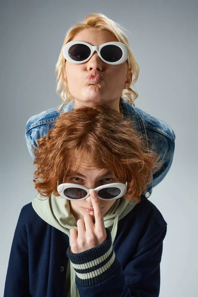 stock image blonde teen girl pouting lips near redhead friend looking at camera over stylish sunglasses on grey