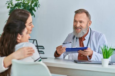 Neşeli gri sakallı doktor kucaklayan lgbt çiftiyle konuşuyor. İçtenlikle gülümsüyorlar.