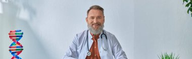 cheerful grey bearded doctor sitting at table looking at camera on bluish backdrop, banner clipart