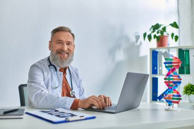 Masasında ultrasonu olan mutlu gri sakallı doktor laptopta çalışıyor ve kameraya samimi bir şekilde gülümsüyor.