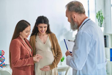 Doktor kontrolündeki neşeli lezbiyen çift hamile karnına, tüp bebek konseptine bakıyor.