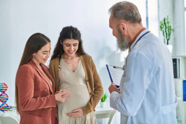 cheerful lesbian couple at doctor checkup looking at pregnant belly, in vitro fertilization concept clipart