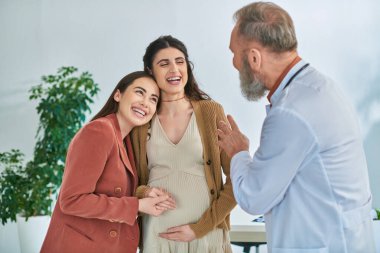 cheerful lgbtq couple hugging warmly and looking at their gynecologist, in vitro fertilizing concept clipart