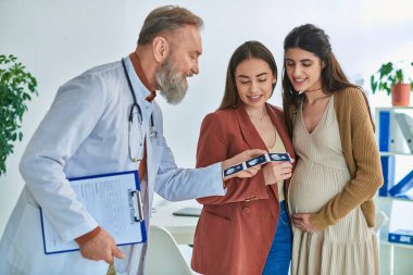 grey bearded doctor showing ultrasound to happy lesbian married couple, in vitro fertilizing concept clipart