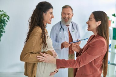 cheerful gynecologist looking at pregnant woman with her partner, in vitro fertilizing concept clipart