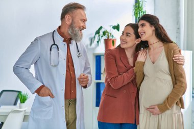 happy lesbian couple hugging with hands on pregnant belly and looking at their doctor, ivf concept clipart