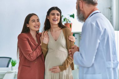 joyous lesbian couple with hands on pregnant belly smiling at their gynecologist, ivf concept clipart