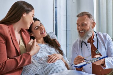jolly doctor showing ultrasound to cheerful smiling lesbian couple, in vitro fertilization concept clipart