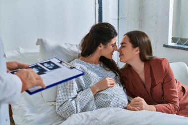 happy lesbian couple rubbing noses at appointment, doctor holding ultrasound in hands, ivf concept clipart