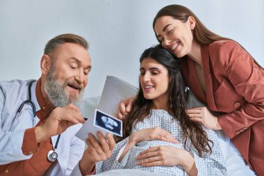 jolly grey bearded doctor showing ultrasound of baby to happy hugging lgbt couple, ivf concept clipart