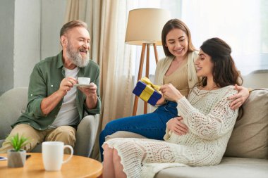 cheerful lesbian couple holding present and father of one of them drinking coffee, ivf concept clipart