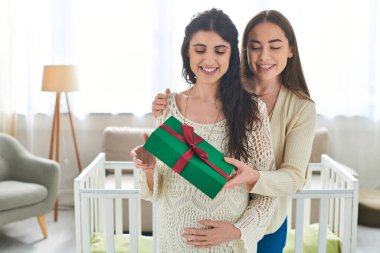 pretty lesbian couple standing next to crib holding present with hand on pregnant belly, ivf concept clipart