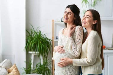 Neşeli lezbiyen çift ellerini hamile karnına koyup sarılıyorlar ve gözlerini kaçırıyorlar.