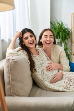 cheerful lgbt couple relaxing on sofa and smiling happily at camera, in vitro fertilisation concept clipart