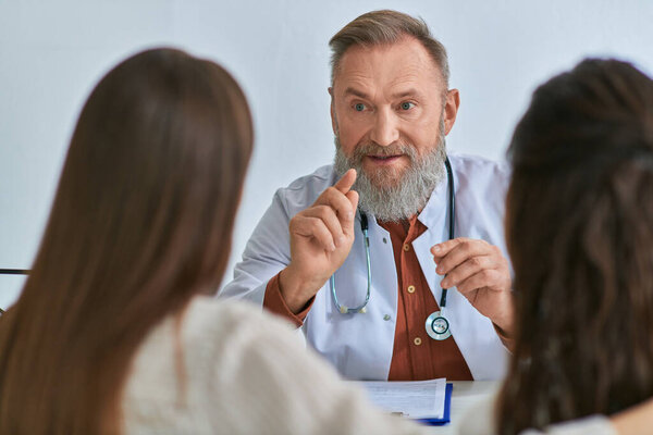 умный веселый доктор жестикулирует и объясняет что-то молодой лесбийской паре, ivf концепция